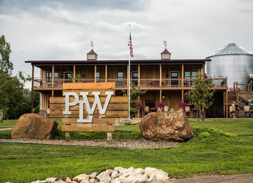 Rustic building with "PLW" sign, wooden facade, stone accents, surrounded by greenery and silos, evoking warmth and tranquility.