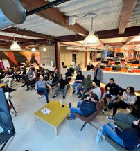 Modern workspace with people engaged in a discussion, featuring varied seating, large windows, and a collaborative atmosphere.