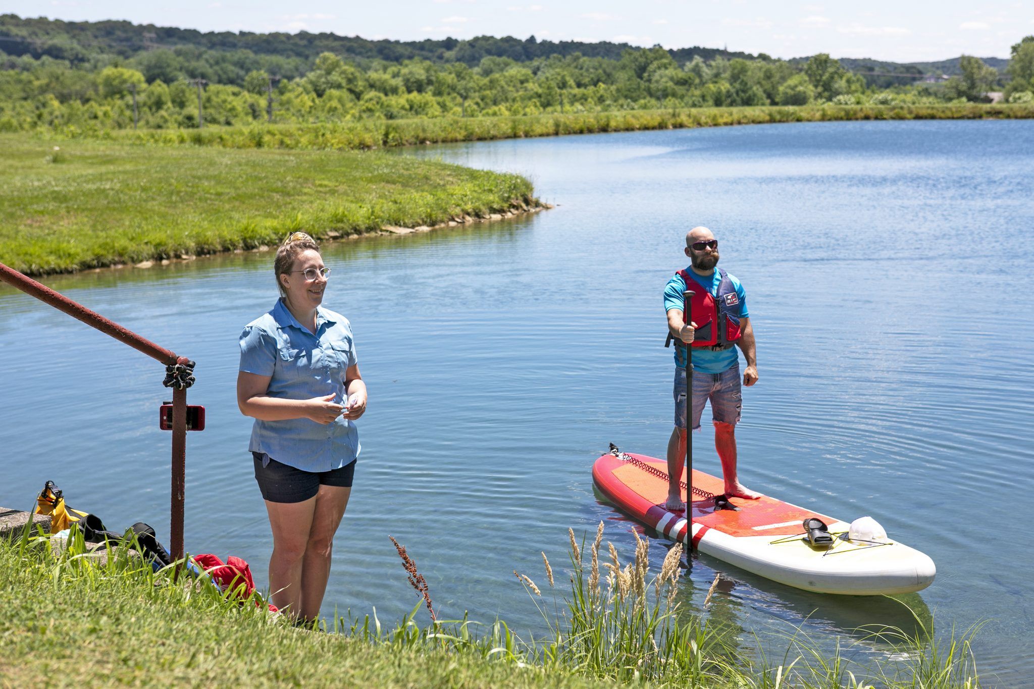 A woman on shore faces a man on a paddleboard by a serene lake, embodying leisure and adventure in a lush, sunny setting.