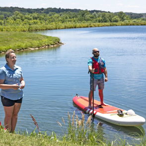 A woman on shore faces a man on a paddleboard by a serene lake, embodying leisure and adventure in a lush, sunny setting.