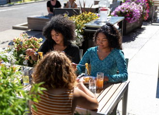 Three people dining outdoors, surrounded by flowers and greenery, enjoying their meal and each other's company in a sunny setting.