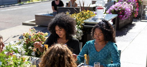 Three people dining outdoors, surrounded by flowers and greenery, enjoying their meal and each other's company in a sunny setting.