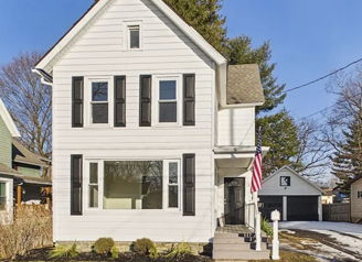 Charming two-story white house with dark shutters, welcoming porch, American flag, green grass, and remnants of snow.