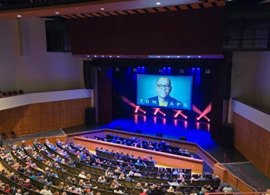A large audience in a tiered auditorium watches a speaker on an illuminated stage, creating an atmosphere of excitement.