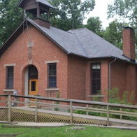 Quaint brick building, likely a former schoolhouse, set in lush greenery, evokes nostalgia and tranquility with its rustic charm.
