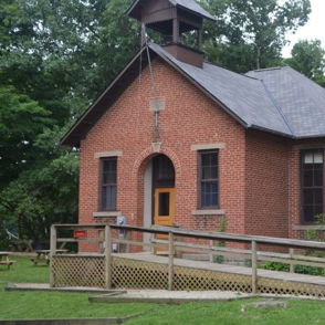 Quaint brick building, likely a former schoolhouse, set in lush greenery, evokes nostalgia and tranquility with its rustic charm.