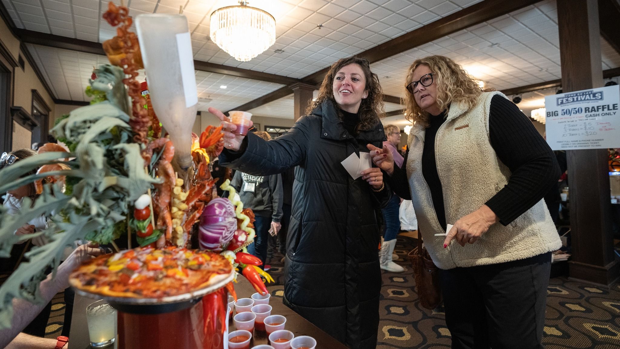 Two women chat joyfully at a food festival, admiring a colorful display of food, evoking excitement and camaraderie.