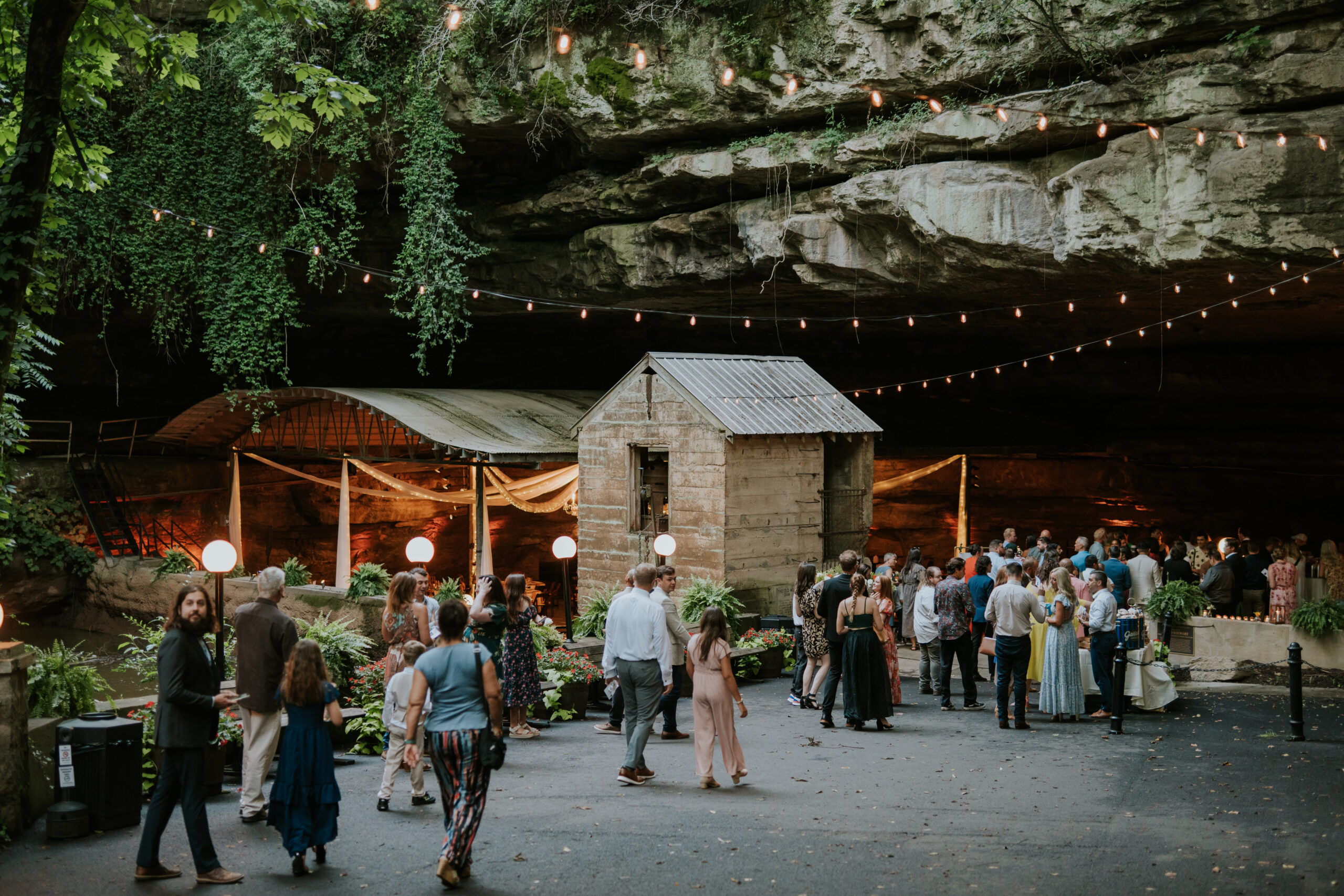 A festive gathering in a cave with string lights, a rustic house, and guests celebrating together, creating a warm atmosphere.