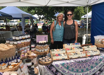 Two smiling women showcase handmade soaps and skincare at a vibrant outdoor market, fostering a cheerful, inviting atmosphere.