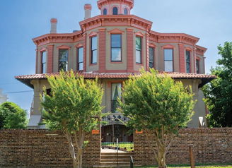 A grand reddish mansion with ornate details, surrounded by lush lawns and a stone wall, evokes nostalgia and tranquility.