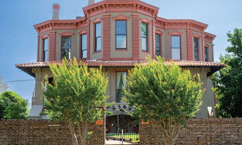 A grand reddish mansion with ornate details, surrounded by lush lawns and a stone wall, evokes nostalgia and tranquility.