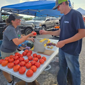 Market scene with a woman in a hat handing a tomato to a man, showcasing community and the joy of local produce.