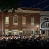An outdoor concert features an orchestra in front of a historic building, lit warmly, with a festive, patriotic atmosphere.