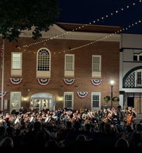 An outdoor concert features an orchestra in front of a historic building, lit warmly, with a festive, patriotic atmosphere.