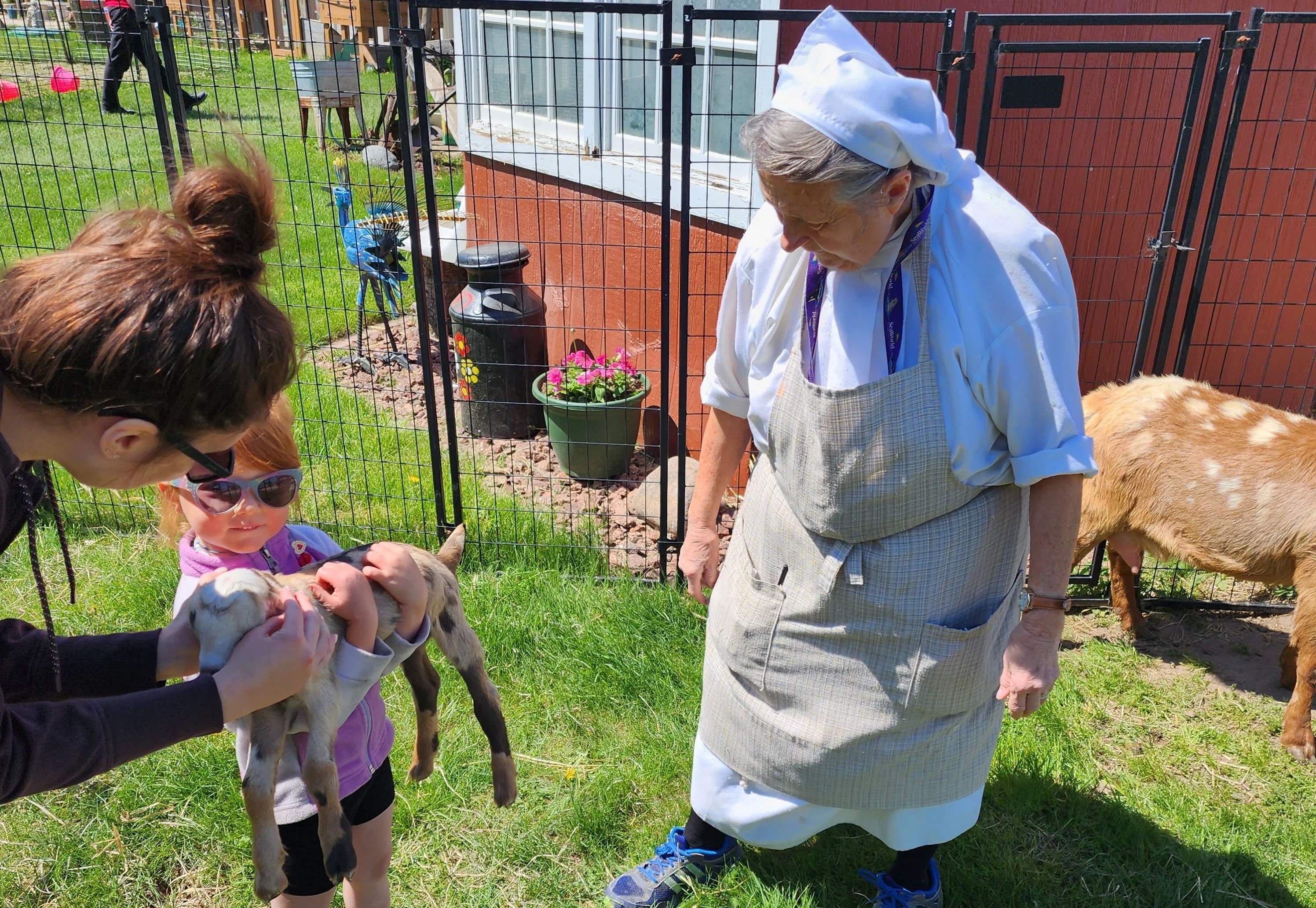 A girl with sunglasses holds a small animal, engaged warmly with an adult and an elderly person in a lively outdoor setting.