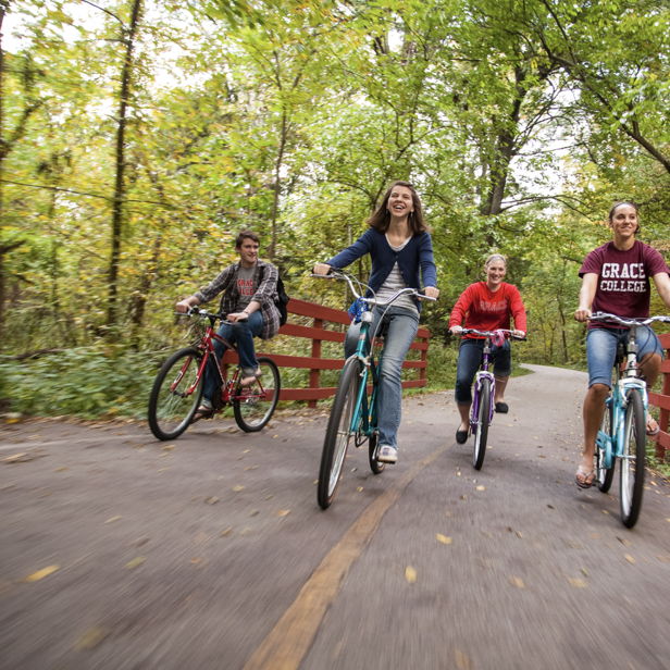 Four happy individuals ride bikes on a vibrant, tree-lined path, enjoying spring/summer and the joy of outdoor adventure.