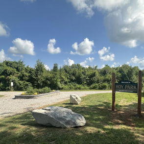 Rhy Park sign in a lush, serene landscape with a gravel path, rocks, blue sky, and fluffy clouds, evoking tranquility.