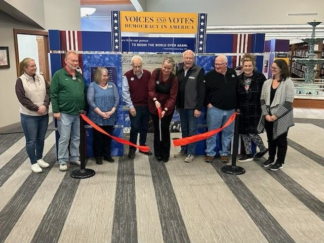 A cheerful group at a ribbon-cutting for a voting-themed event titled "Voices and Votes," evoking community spirit and pride.