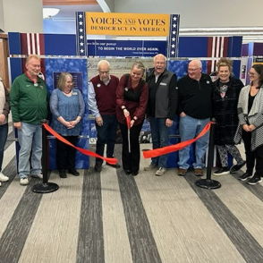 A cheerful group at a ribbon-cutting for a voting-themed event titled "Voices and Votes," evoking community spirit and pride.
