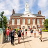 Group of people outside a sunny school building with a clock tower, engaged in conversation, evoking community and enthusiasm.