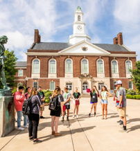 Group of people outside a sunny school building with a clock tower, engaged in conversation, evoking community and enthusiasm.