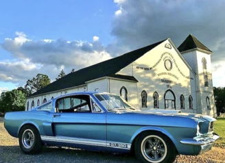 Classic blue car parked by a grand white building, with fluffy clouds above, evoking nostalgia and a sense of adventure.