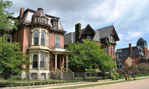 Row of detailed historical houses with Victorian styles, lush greenery, and an overcast sky, evoking nostalgia and history.