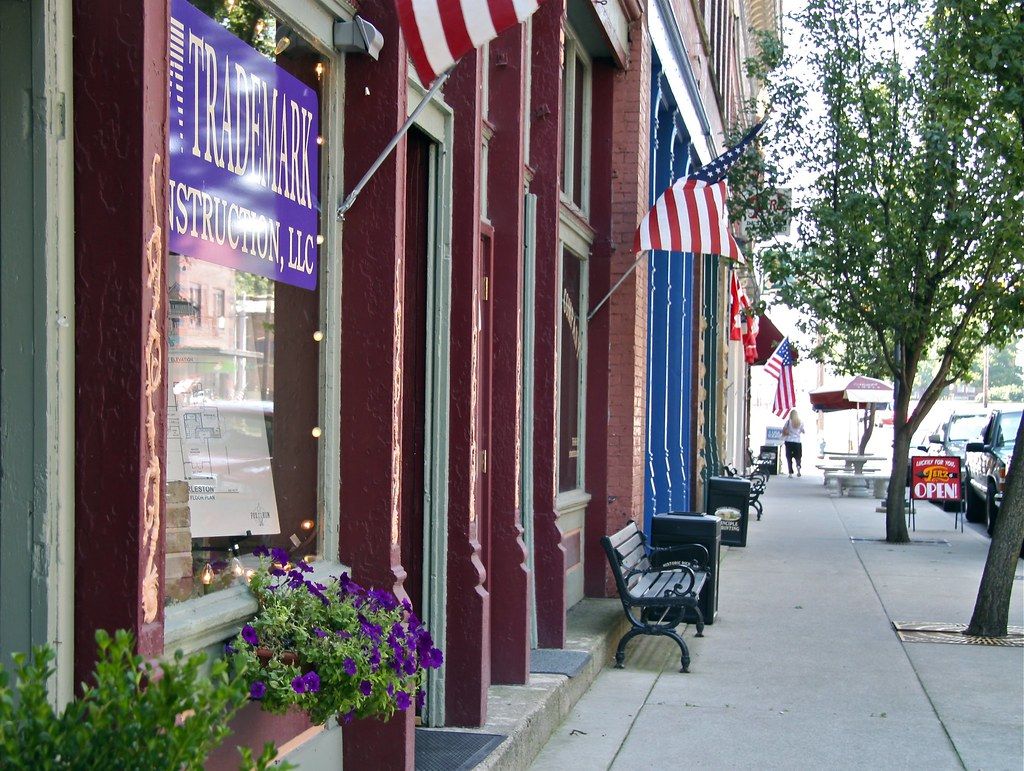 Charming street scene with trees, benches, American flags, vibrant storefront, purple flowers; evoking community and nostalgia.