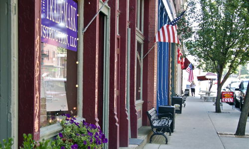 Charming street scene with trees, benches, American flags, vibrant storefront, purple flowers; evoking community and nostalgia.