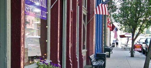 Charming street scene with trees, benches, American flags, vibrant storefront, purple flowers; evoking community and nostalgia.