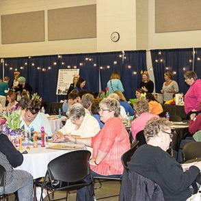 Lively gathering of mostly women at a community event, engaging in crafts with colorful decorations and a festive atmosphere.