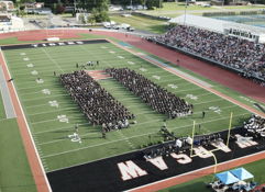 Graduation ceremony on a sports field with graduates in black gowns, surrounded by a supportive crowd, celebrating achievement.