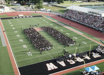 Graduation ceremony on a sports field with graduates in black gowns, surrounded by a supportive crowd, celebrating achievement.