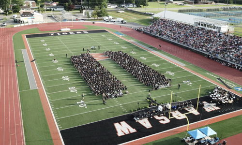 Graduation ceremony on a sports field with graduates in black gowns, surrounded by a supportive crowd, celebrating achievement.