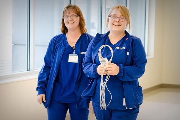 Two smiling healthcare professionals in blue scrubs hold medical equipment in a bright, clean corridor, evoking warmth and trust.