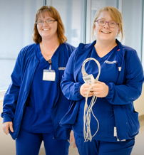 Two smiling healthcare professionals in blue scrubs hold medical equipment in a bright, clean corridor, evoking warmth and trust.
