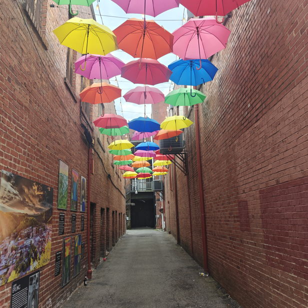 A vibrant alleyway with colorful umbrellas overhead and exposed brick walls creates a cheerful, whimsical atmosphere.