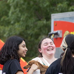 Young women joyfully interacting outdoors, one holding a dog, sharing laughter and friendship in a vibrant, festive atmosphere.