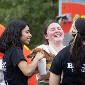 Young women joyfully interacting outdoors, one holding a dog, sharing laughter and friendship in a vibrant, festive atmosphere.