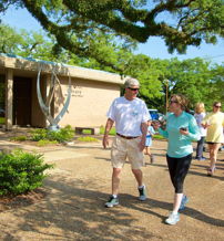 A lively scene of people walking outside, enjoying a sunny day with greenery and a sculpture, radiating joy and camaraderie.