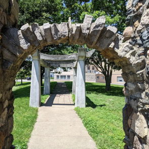 Stone archway leads to a serene park pathway, framed by vibrant greenery, evoking tranquility and a sense of adventure.