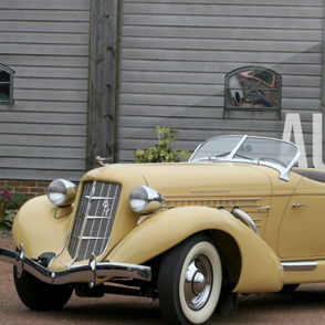 Vintage Auburn car in soft yellow, convertible top, against rustic building, evokes nostalgia and admiration for classic design.