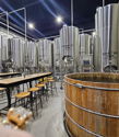 Industrial brewing facility with stainless steel tanks, rustic wooden barrel, long table, and cozy lighting for gatherings.