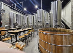 Industrial brewing facility with stainless steel tanks, rustic wooden barrel, long table, and cozy lighting for gatherings.
