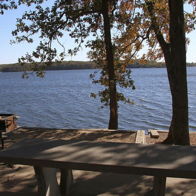A serene lakeside scene with a picnic table under trees, autumn leaves, a grill, and a calm, inviting atmosphere.