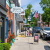 Charming street scene with shops, greenery, American flags, and people enjoying a sunny day, evoking warmth and community.