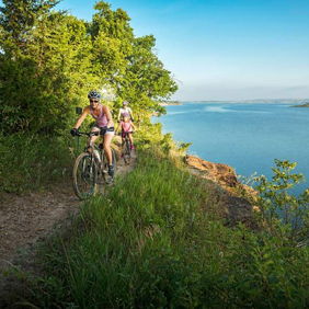 Two individuals bike along a scenic trail by water, surrounded by greenery, evoking adventure and tranquility in nature.