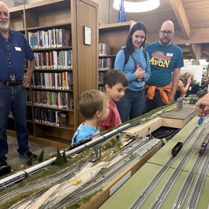 A library scene with children fascinated by a model train setup, while an adult demonstrates, creating joy and curiosity.