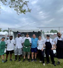 A group in casual and chef attire stands by a tent, showcasing camaraderie and excitement at a food festival under cloudy skies.