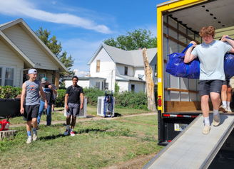 A group of young people collaboratively unload a truck on a sunny day, showcasing teamwork and a sense of new beginnings.